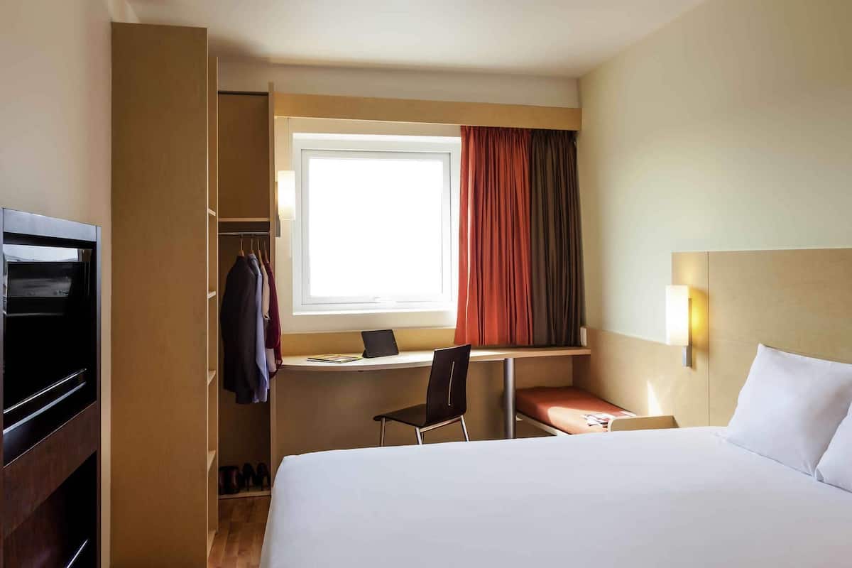 A hotel room with a bed, a desk with a chair, a wardrobe, and a window with curtains.