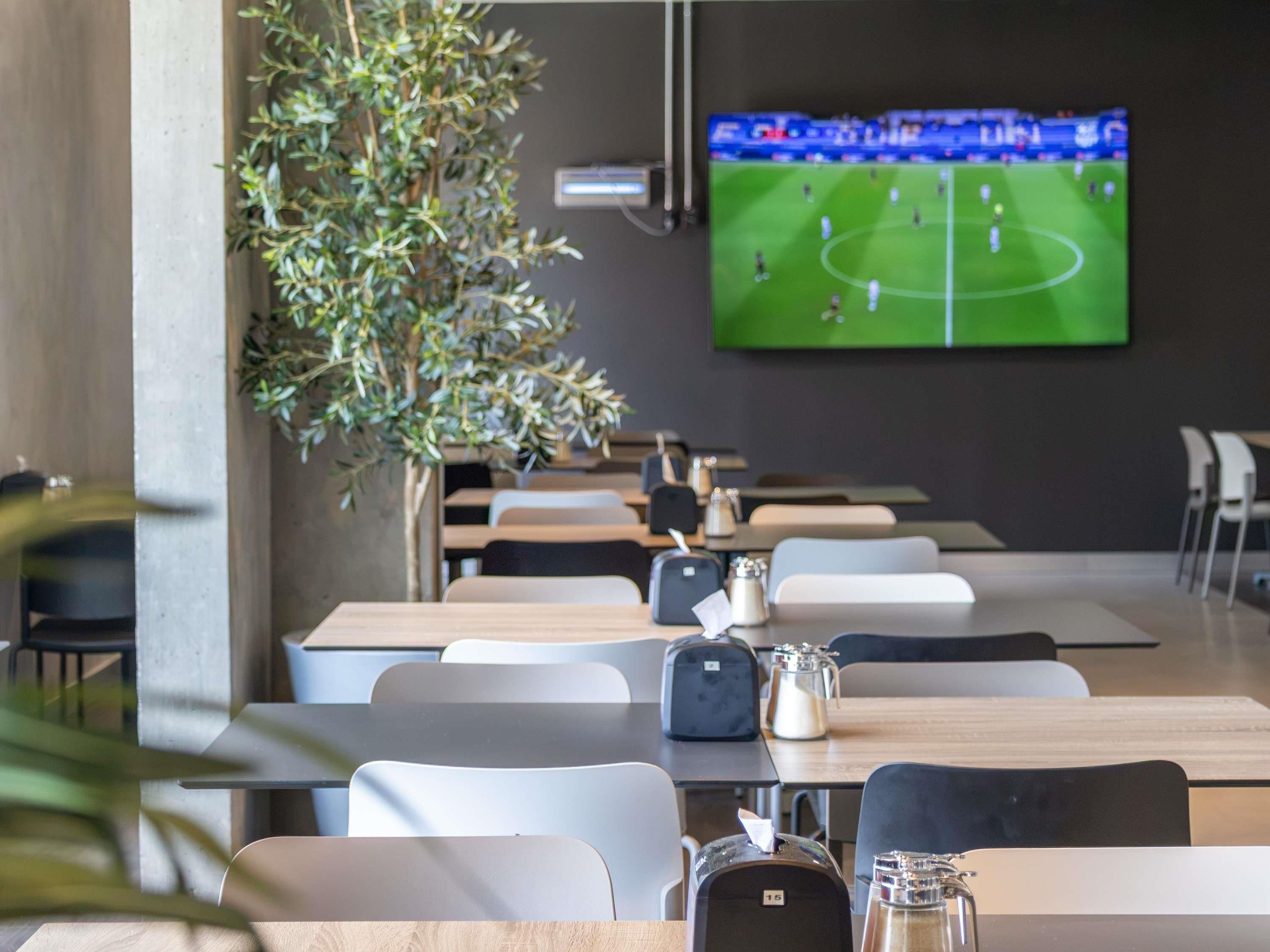 Un comedor moderno con mesas y sillas, un televisor mostrando un partido de fútbol y una planta en maceta.