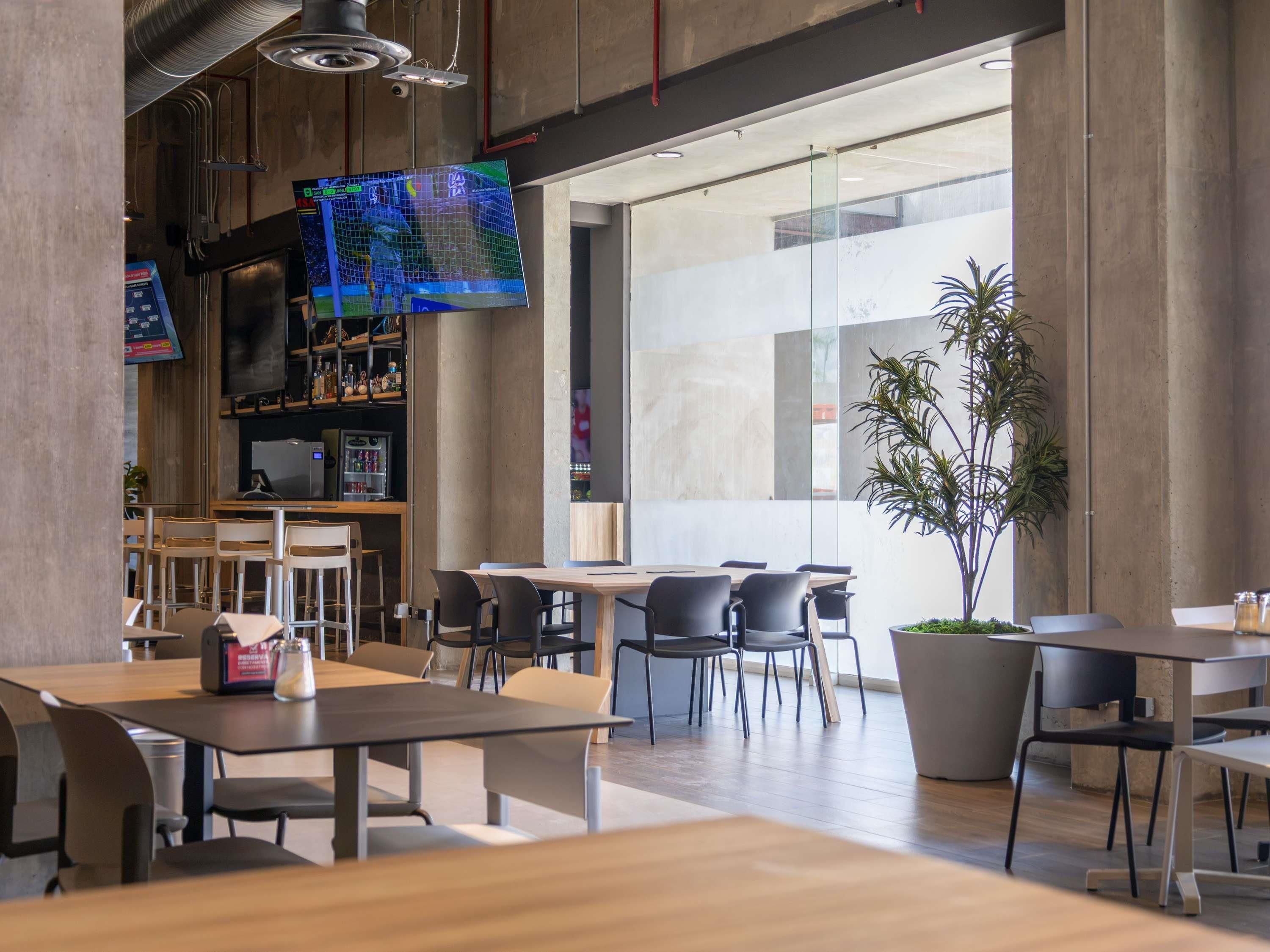 Una cafetería moderna con mesas, sillas, una pantalla de televisión y una planta grande en maceta.