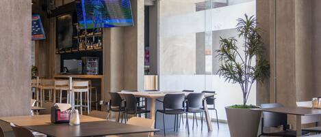 Una cafetería moderna con mesas, sillas, una pantalla de televisión y una planta grande en maceta.