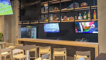 Zona de bar con sillas y mesas de madera, estanterías con botellas y recuerdos deportivos, y una pantalla de televisión mostrando un evento deportivo.