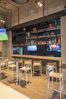 Zona de bar con sillas y mesas de madera, estanterías con botellas y recuerdos deportivos, y una pantalla de televisión mostrando un evento deportivo.
