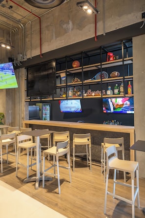 A bar area with wooden chairs and tables, shelves with bottles and sports memorabilia, and a television screen displaying a sports event.
