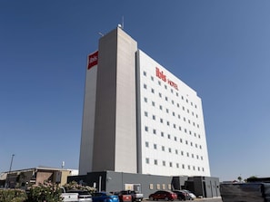 A tall hotel building with a prominent 'ibis hotel' sign, multiple windows, and a parking area with several cars.