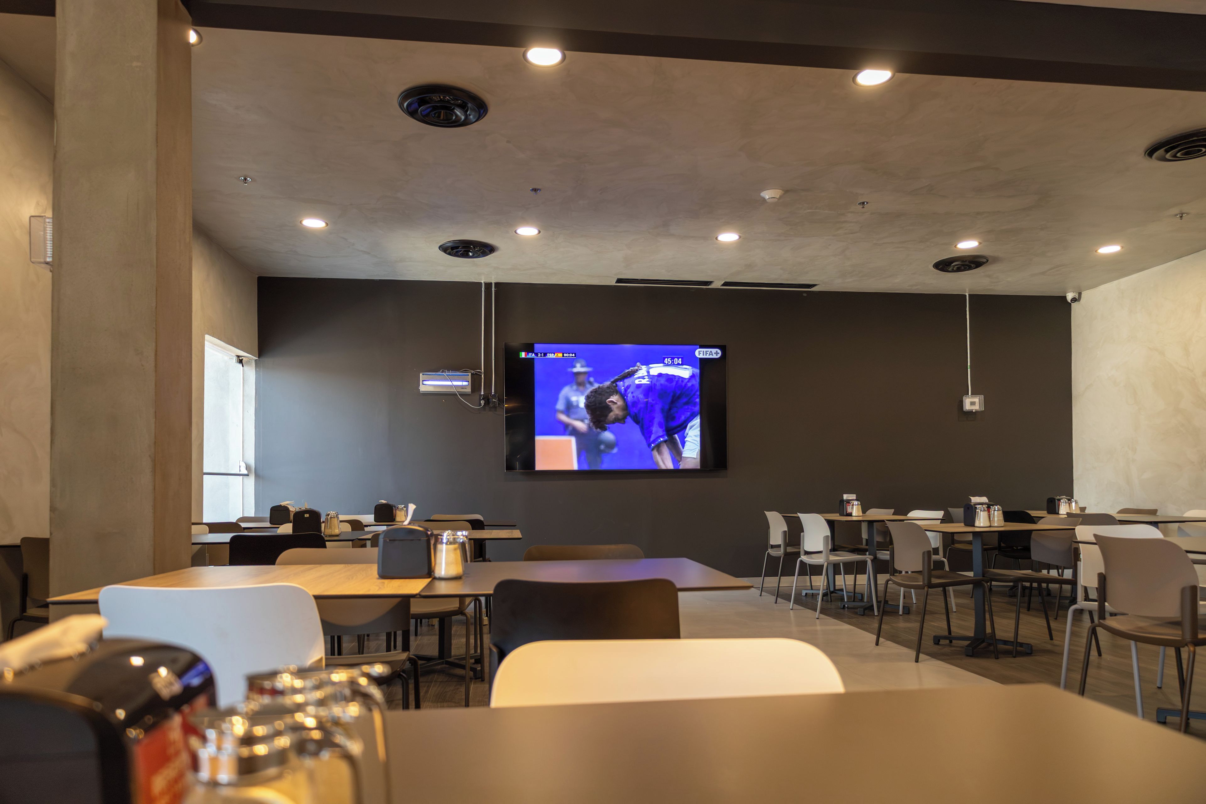Un salón de conferencias con una pantalla grande mostrando un evento deportivo, varias mesas y sillas dispuestas para reuniones.