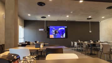 Un salón de conferencias con una pantalla grande mostrando un evento deportivo, varias mesas y sillas dispuestas para reuniones.