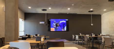 Un salón de conferencias con una pantalla grande mostrando un evento deportivo, varias mesas y sillas dispuestas para reuniones.