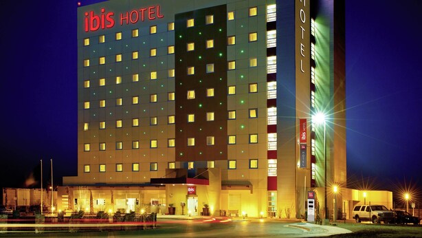 Un hotel de varios pisos con un llamativo letrero 'ibis HOTEL', una entrada espaciosa y un área de estacionamiento.
