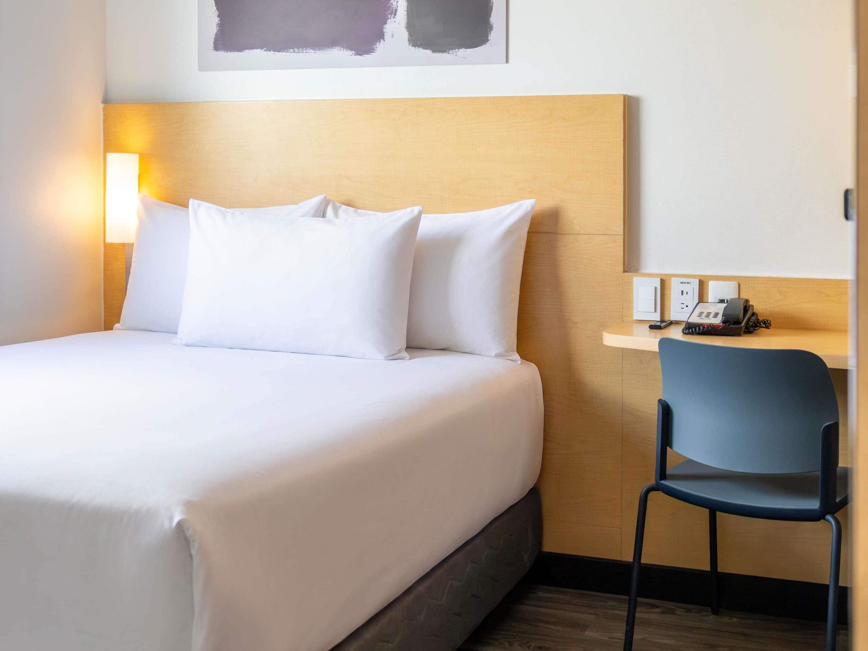 A hotel room with a bed, pillows, a desk with a chair, a phone, and a wall-mounted light.