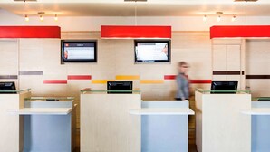 Un área de recepción de hotel moderna con tres mostradores de vidrio, dos pantallas digitales mostrando información y un piso de baldosas limpias.