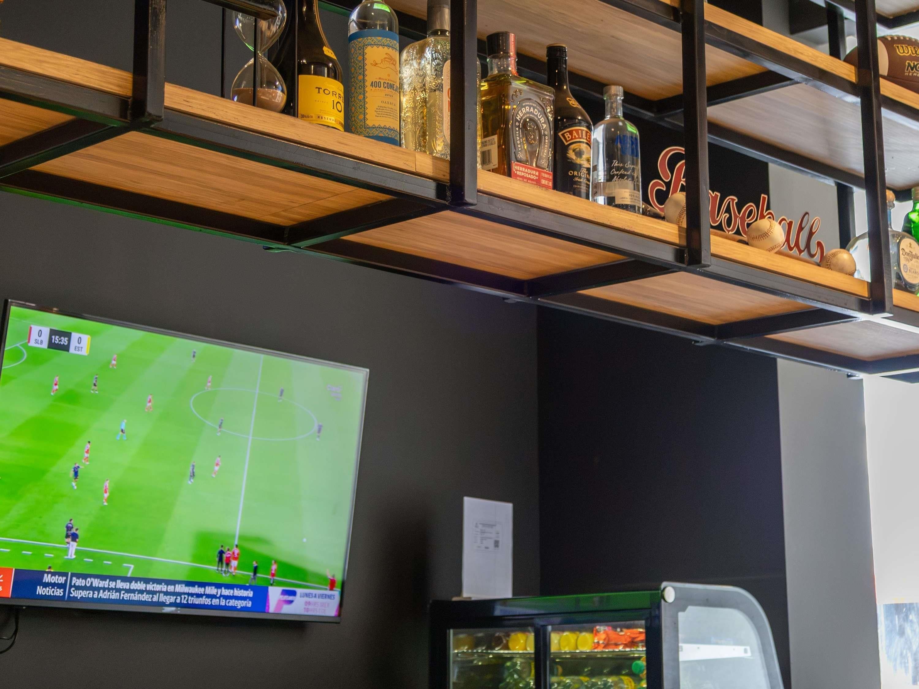 A bar area with shelves holding liquor bottles and a television displaying a soccer match.
