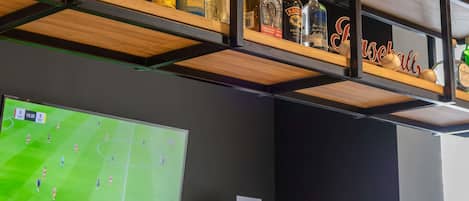 Zona de bar con estantes que exhiben botellas de licor y un televisor mostrando un partido de fútbol.