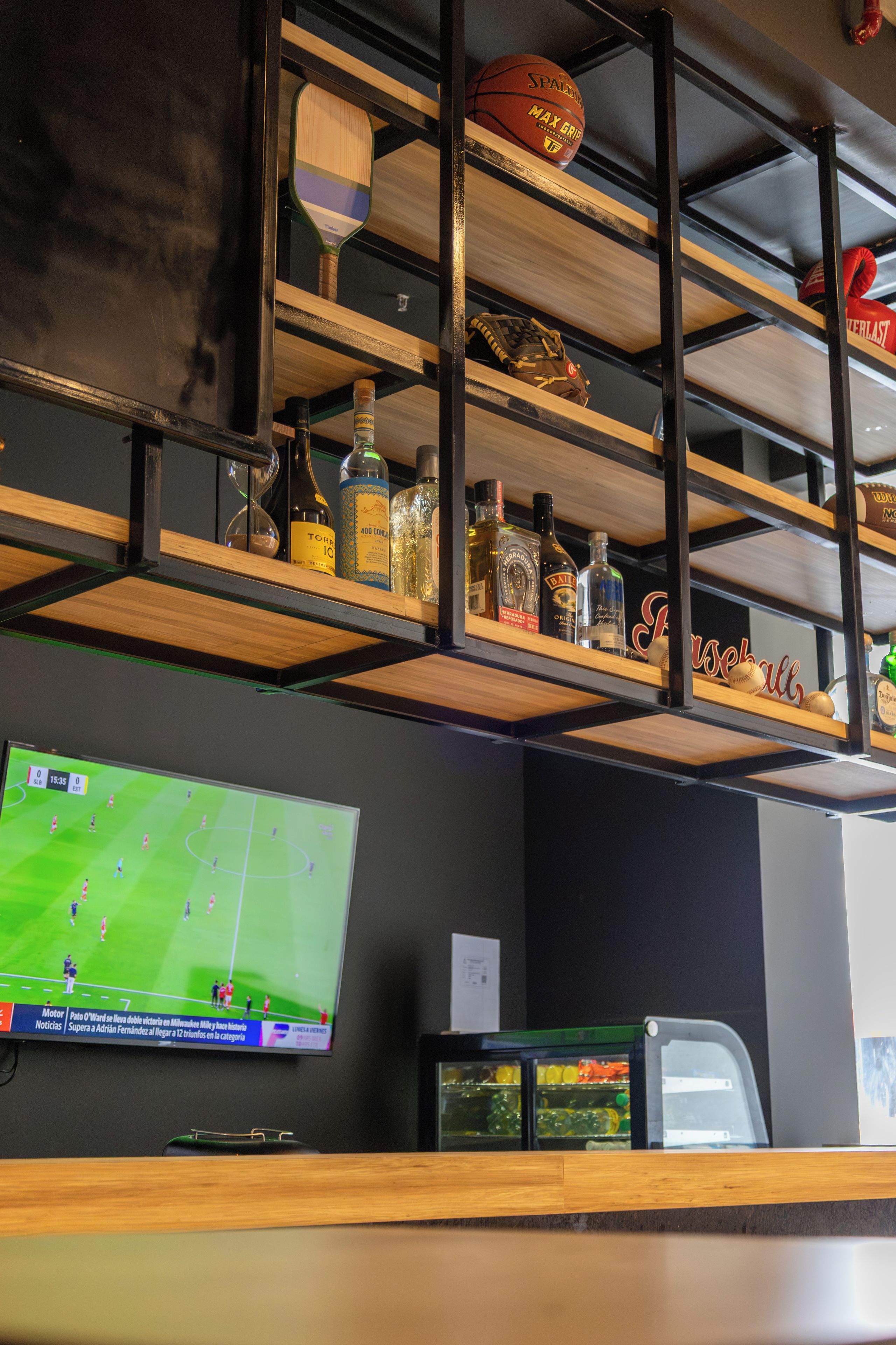 A bar area with shelves holding bottles, a sports ball, and a television displaying a soccer game.