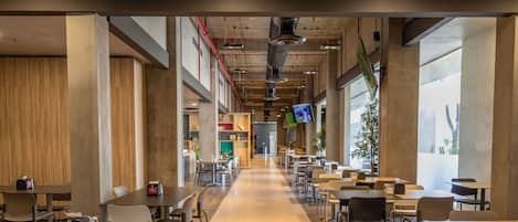 A spacious indoor area with multiple tables and chairs arranged for dining, large windows allowing natural light, and a visible kitchen area in the background.