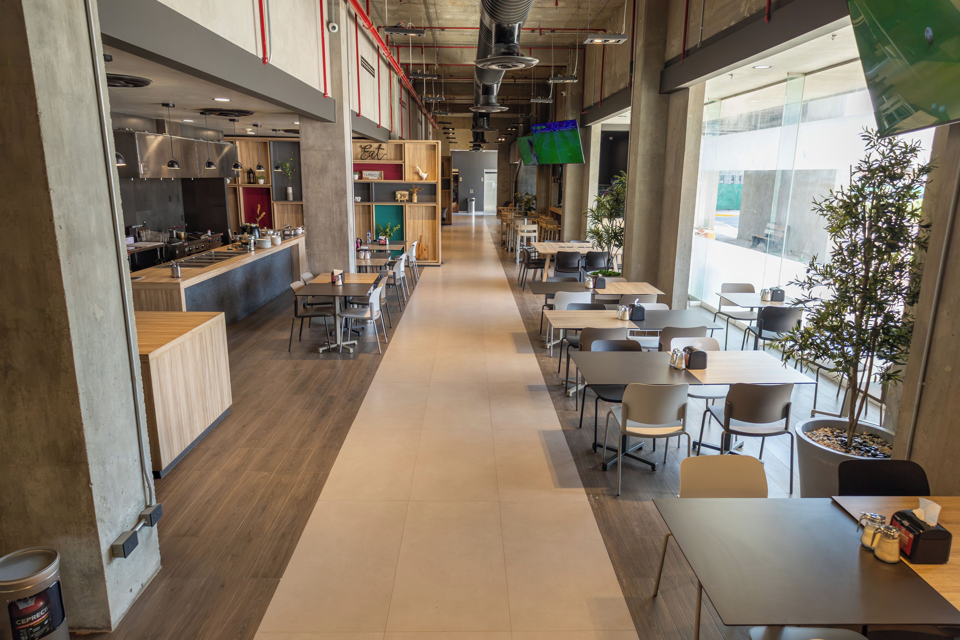 Una amplia cafetería con numerosas mesas y sillas, una zona de cocina y grandes ventanales que dejan entrar luz natural.