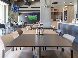 A modern dining area with a long table, multiple chairs, a television screen, and a kitchen counter.