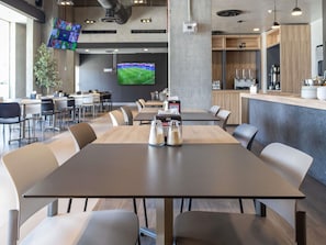 A modern dining area with a long table, multiple chairs, a television screen, and a kitchen counter.