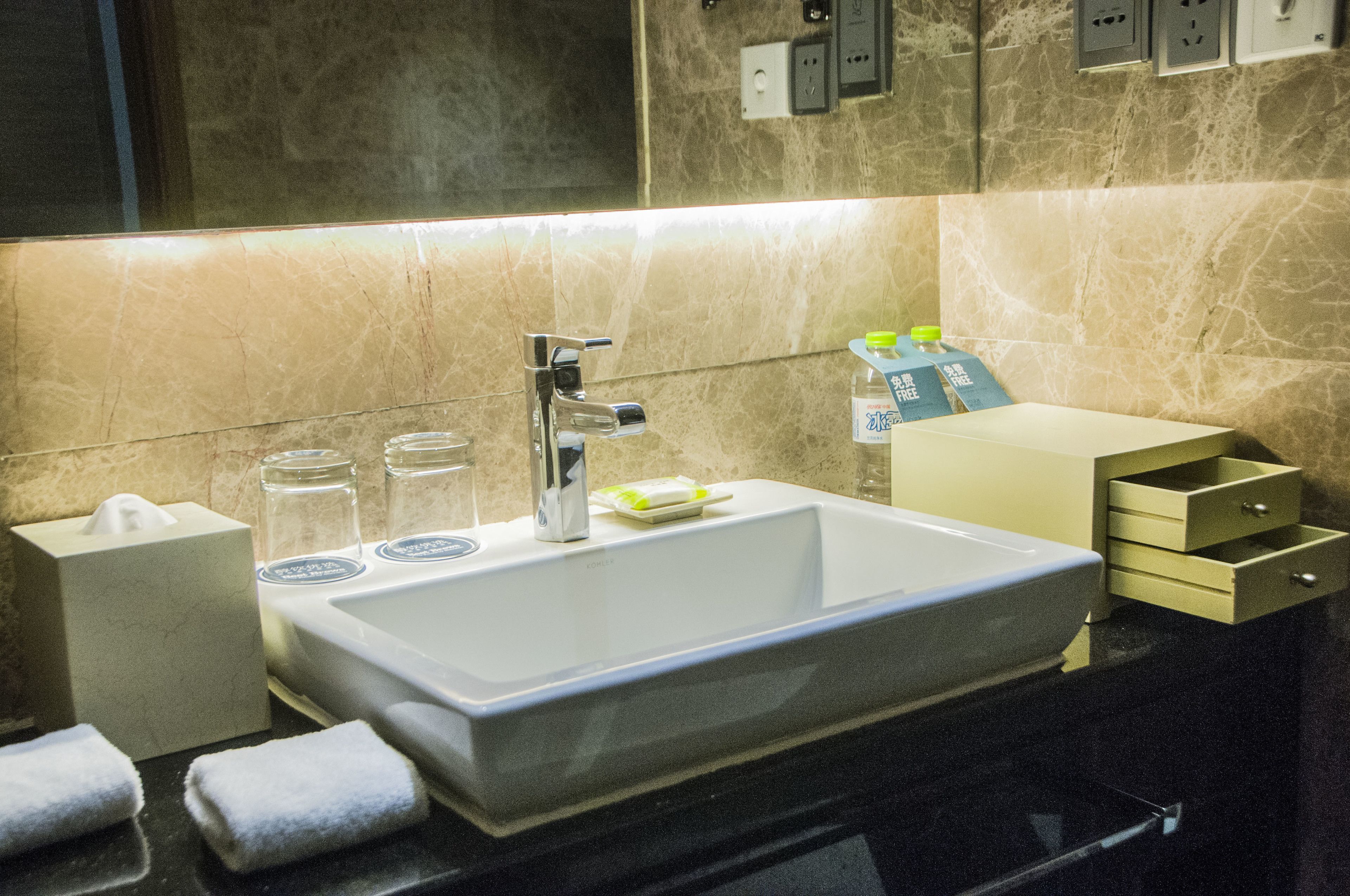 A modern bathroom with a large sink, a mirror, and a towel rack. There are toiletries on the counter and a small cabinet to the right.