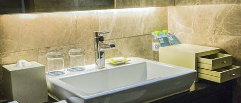 A modern bathroom with a large sink, a mirror, and a towel rack. There are toiletries on the counter and a small cabinet to the right.
