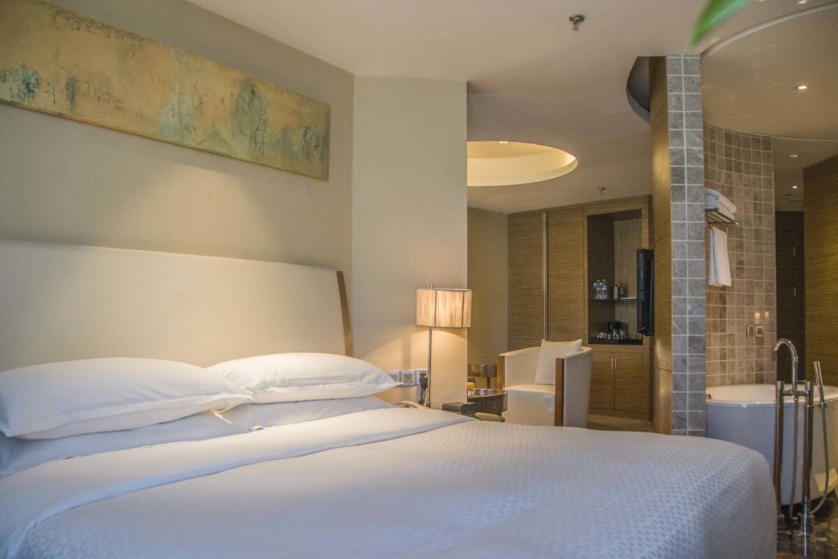 Habitación de hotel con una cama grande, una mesita de noche, una lámpara, baño con ducha y un cuadro en la pared.