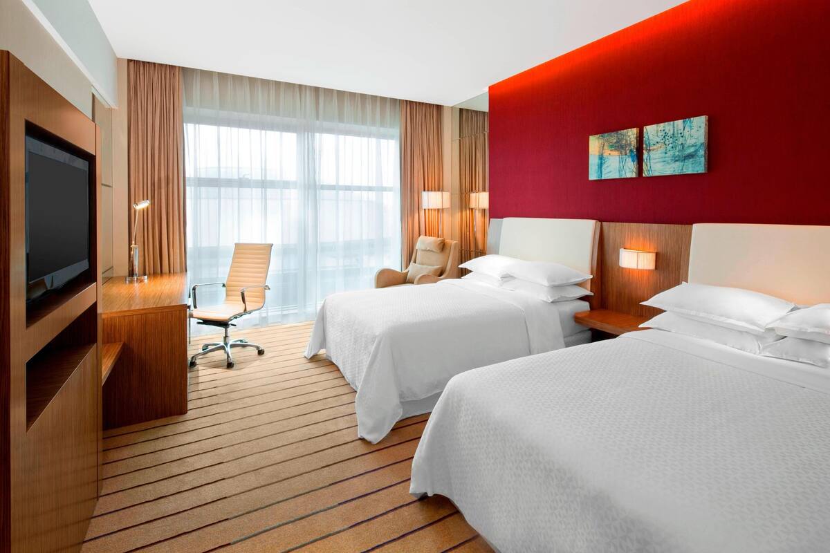 Habitación de hotel con dos camas, un escritorio con televisor, una silla y un ventanal con cortinas.