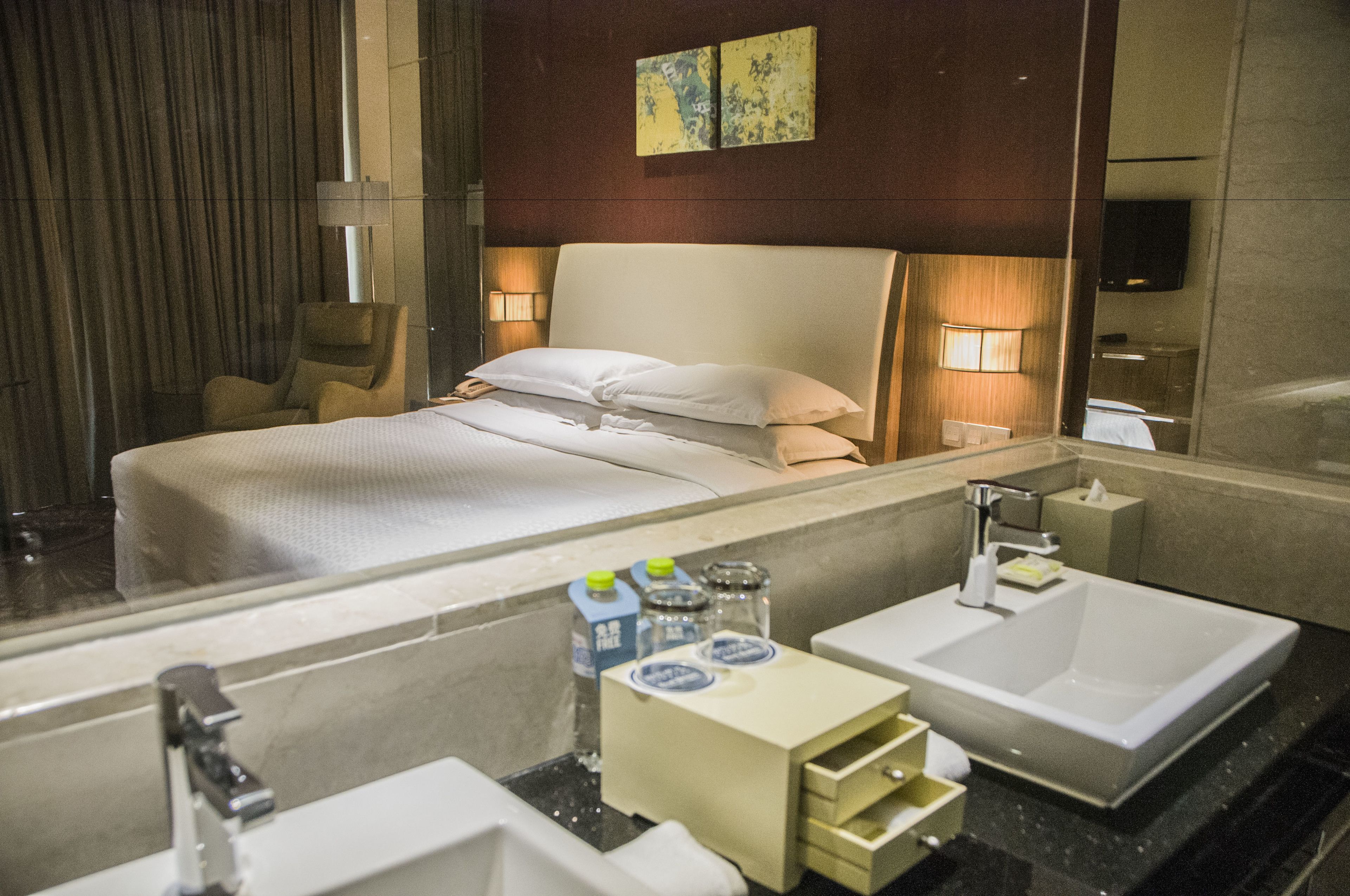 A hotel room with a large bed, a sitting area, a bathroom with a sink and mirror, and a flat-screen TV mounted on the wall.