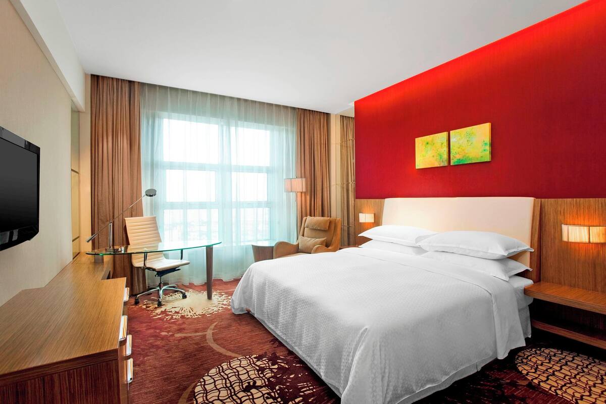 Habitación de hotel con una cama grande, un escritorio con una silla, un televisor y una ventana con cortinas.