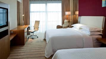 A hotel room with two beds, a desk, a chair, a TV, and large windows.