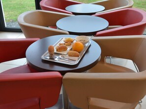 Un espace détente avec des tables et des chaises rondes, un plateau contenant de la nourriture et des boissons, et une vue sur l’extérieur par la fenêtre.