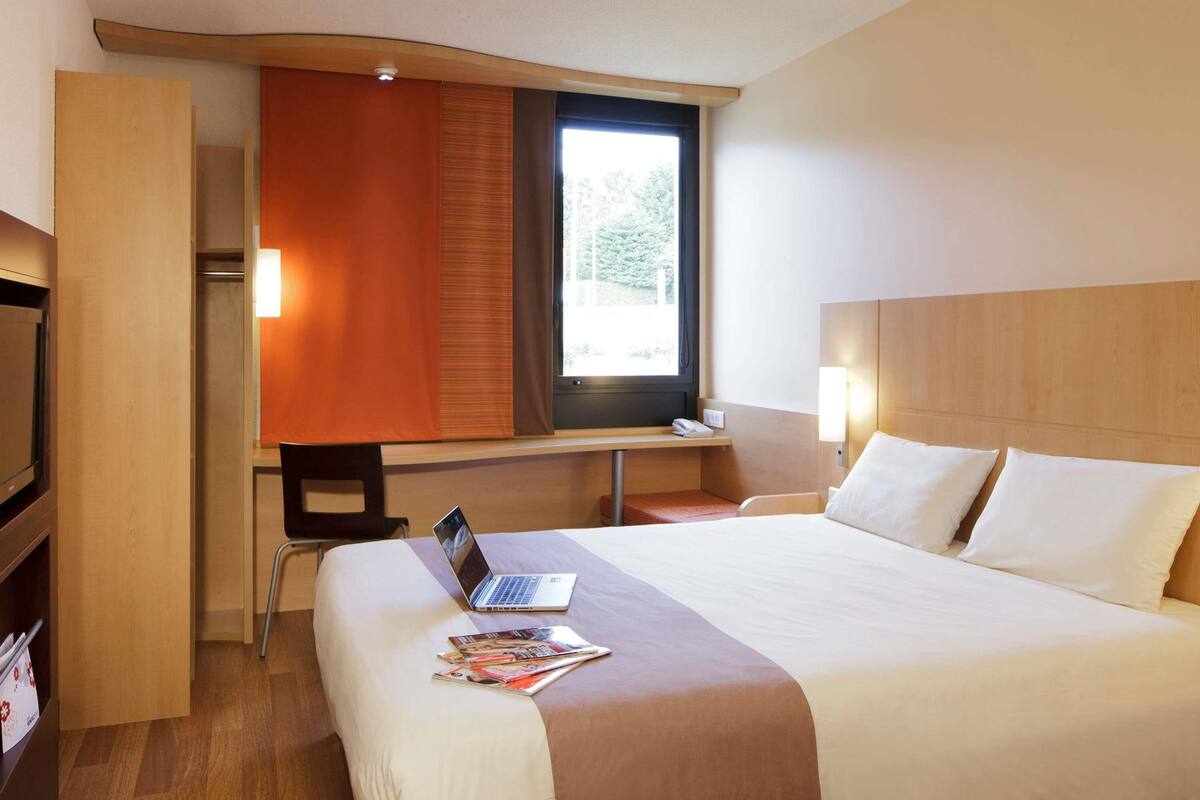 A hotel room with a bed, a desk with a laptop, a chair, a TV, and a window with blinds.
