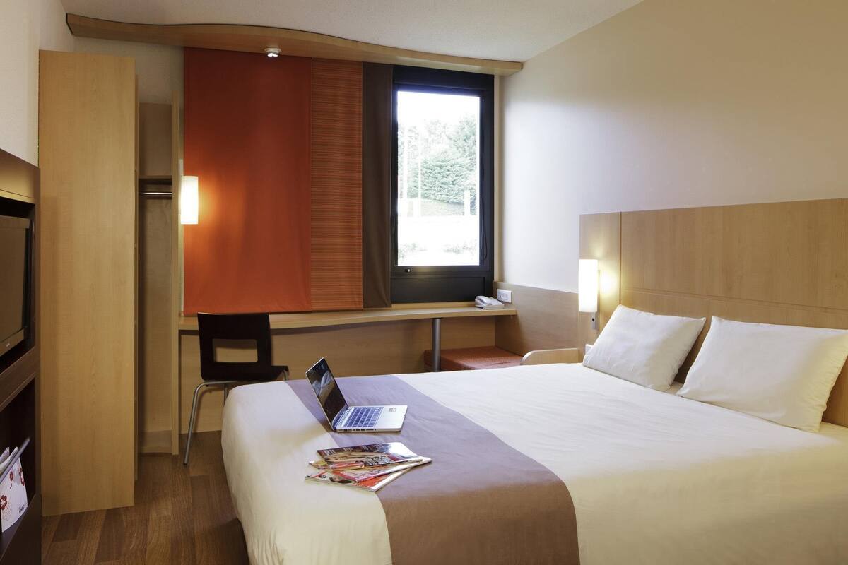 A hotel room with a bed, a desk with a laptop, a TV, and a window with curtains.