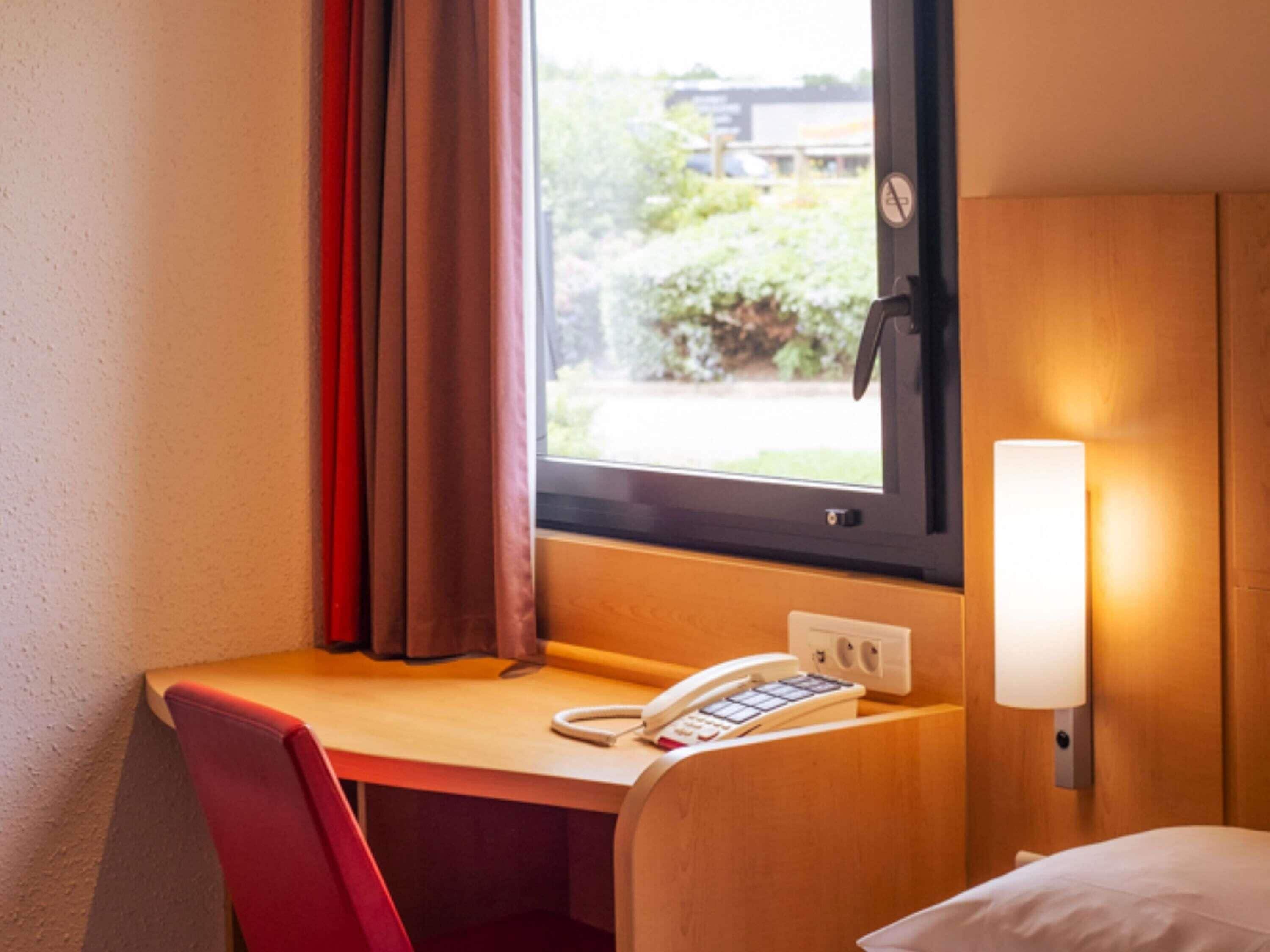 Habitación de hotel con escritorio de madera, teléfono, lámpara, ventana con cortinas y una cama.