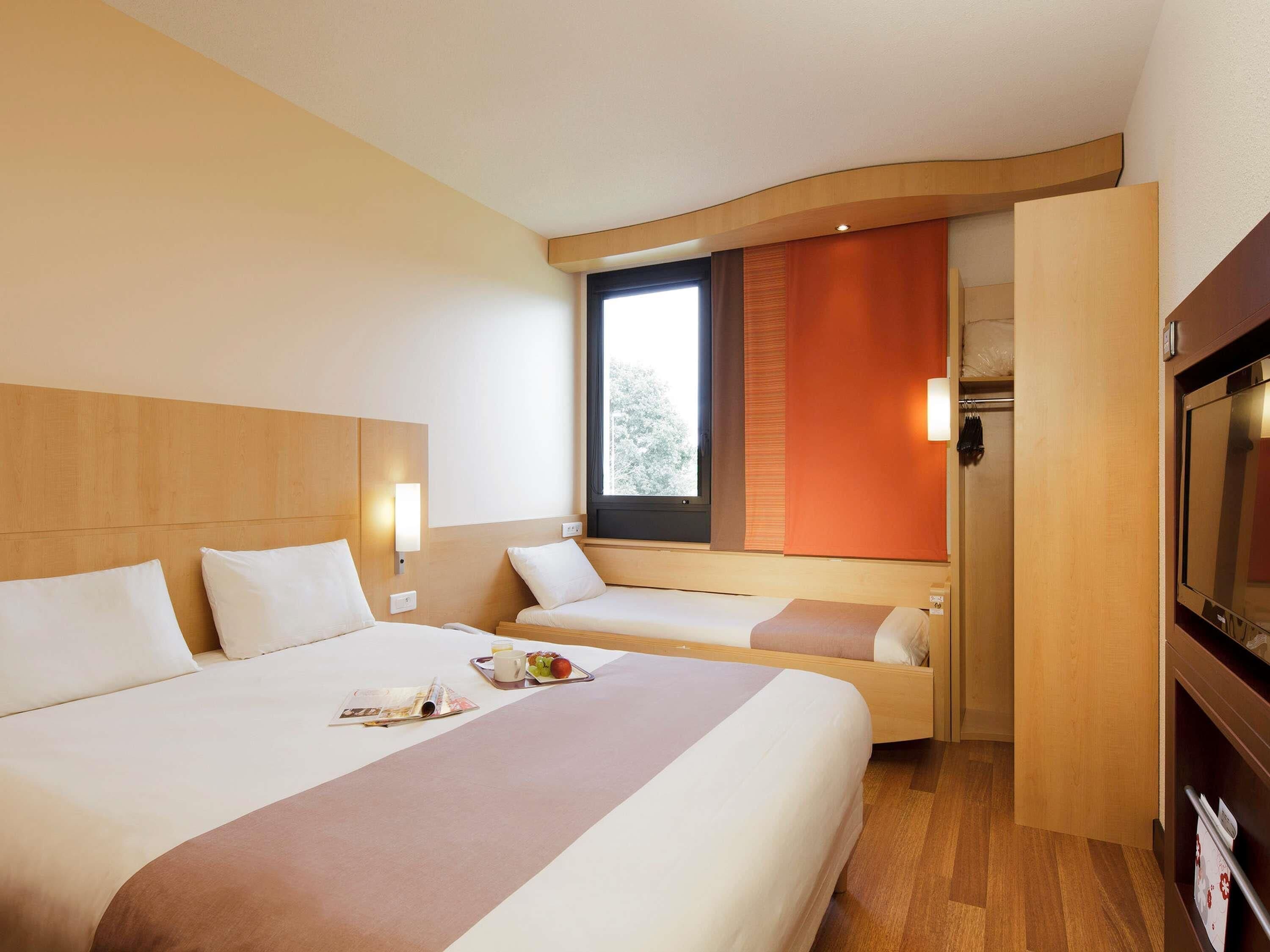 Ein Hotelzimmer mit einem großen Bett, einem kleineren Bett, einem Fernseher und einem Fenster mit Blick auf Bäume.