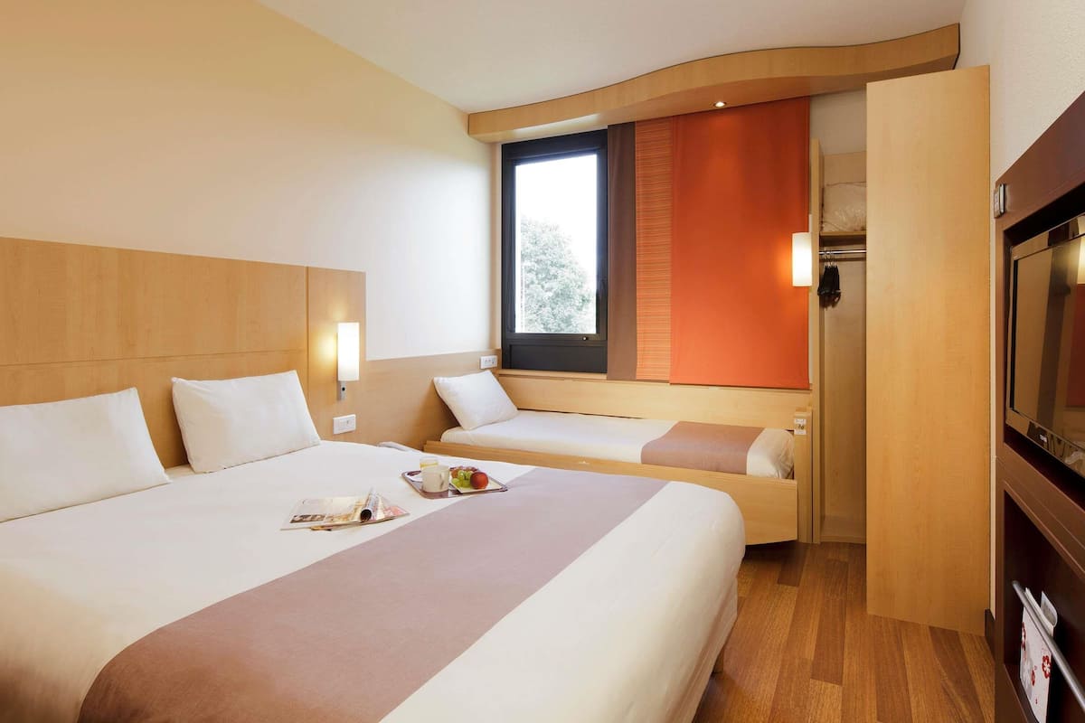 Habitación de hotel con una cama grande, una cama más pequeña, un televisor y una ventana con vistas a árboles.