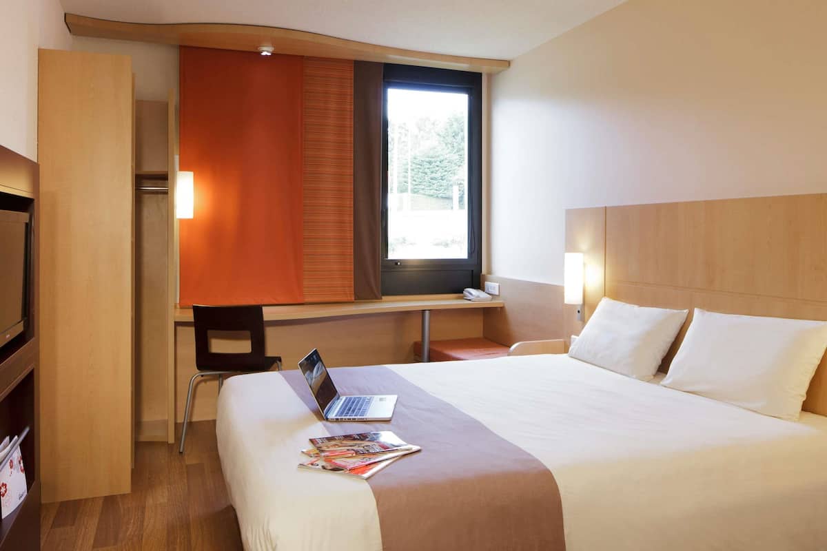 Habitación de hotel con cama, escritorio con ordenador portátil, silla, televisor y ventana con persianas.