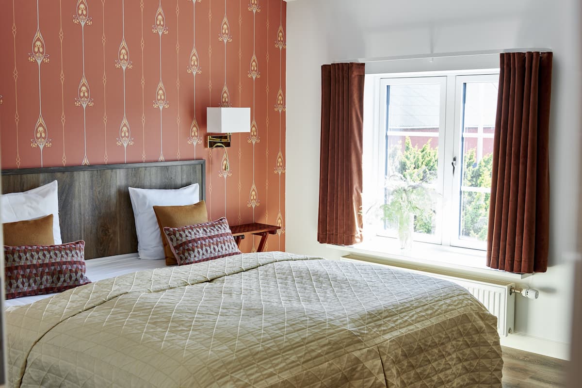Una habitación de hotel con una cama grande, una mesita de noche, una ventana con cortinas y una pared con papel tapiz estampado.