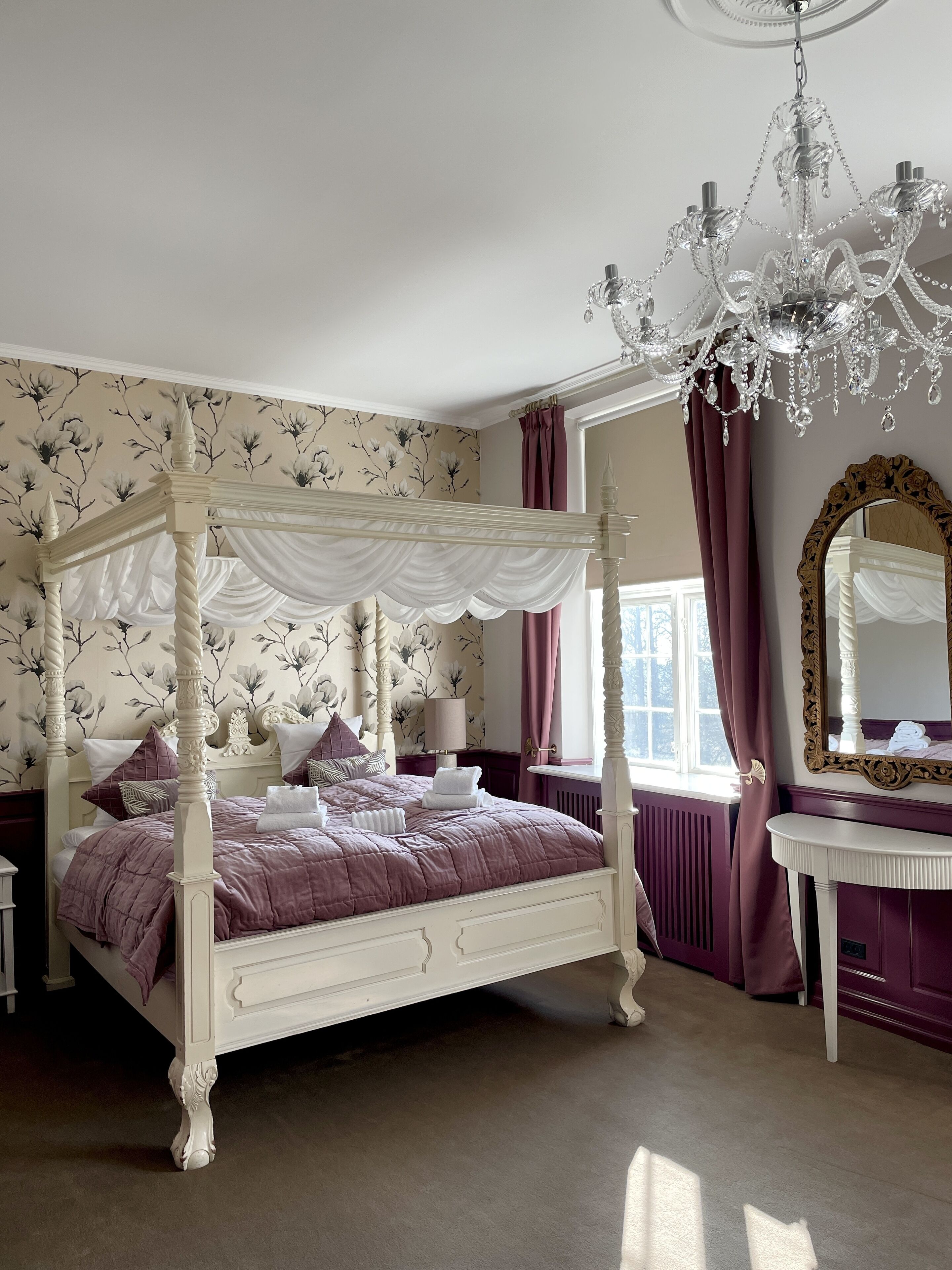 A four-poster bed with a canopy, a chandelier, a vanity with a mirror, and floral wallpaper.