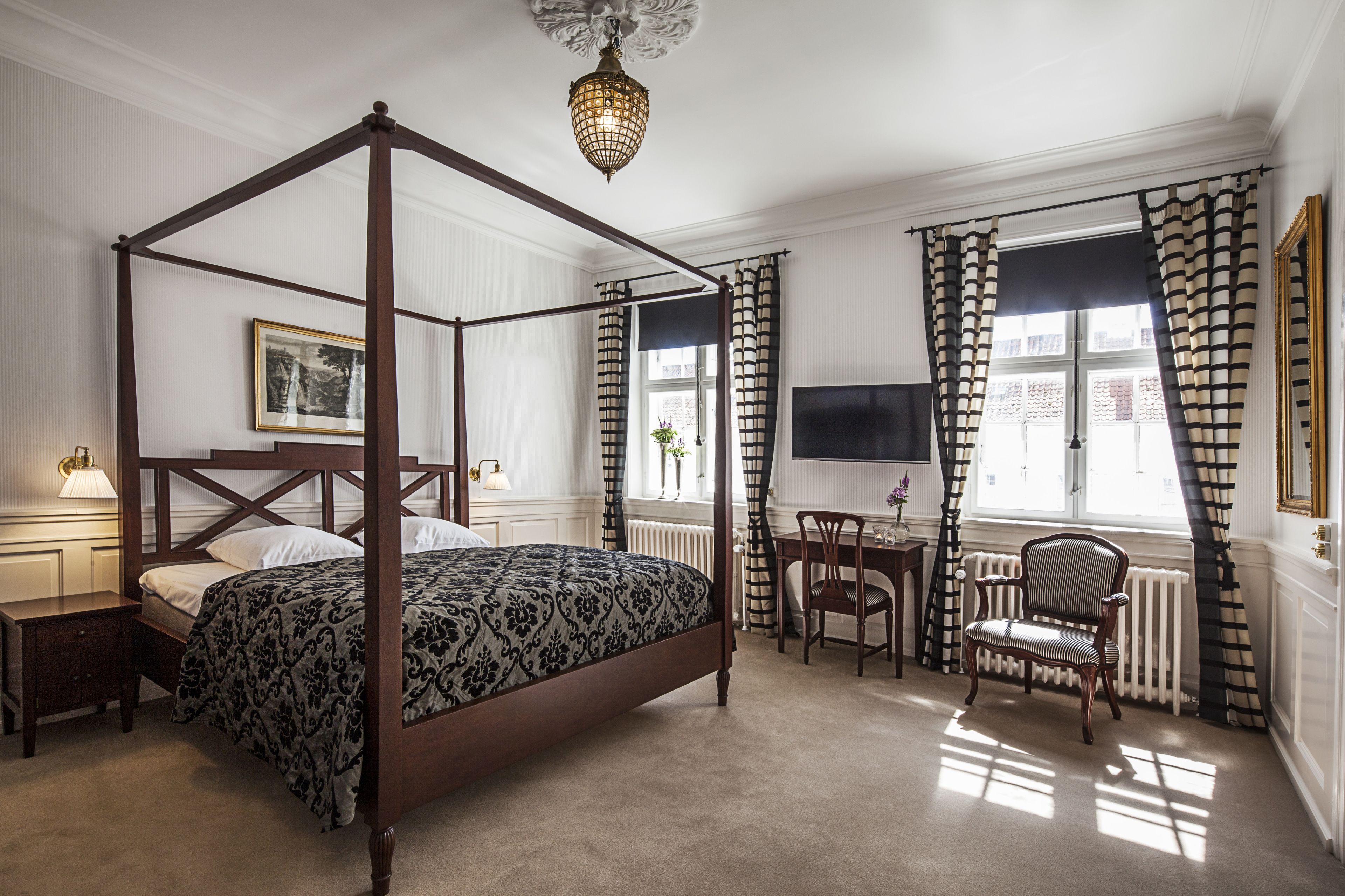 Een hemelbed met een donker houten frame en een beddegoed met een patroon, een klein nachtkastje met een lamp, een stoel, en een bureau met een stoel, alles in een kamer met witte wanden en een groot raam met gestreepte gordijnen.