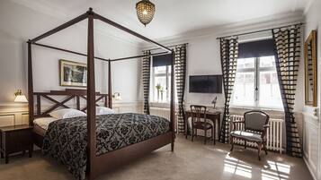 Een hemelbed met een donker houten frame en een beddegoed met een patroon, een klein nachtkastje met een lamp, een stoel, en een bureau met een stoel, alles in een kamer met witte wanden en een groot raam met gestreepte gordijnen.