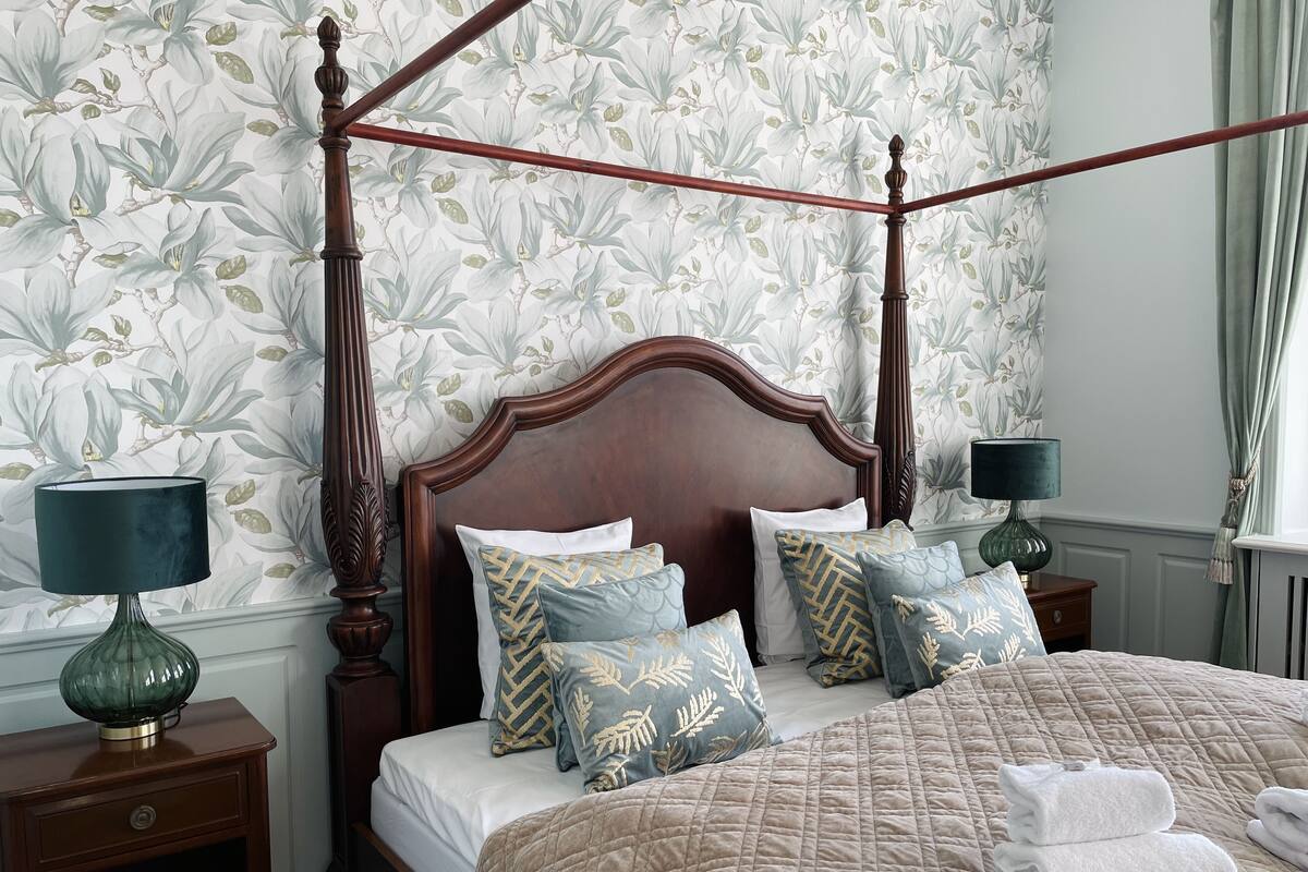 Una cama con dosel y tablero de madera, con varios almohadones, cubrecama acolchado y dos mesitas de noche con lámparas.