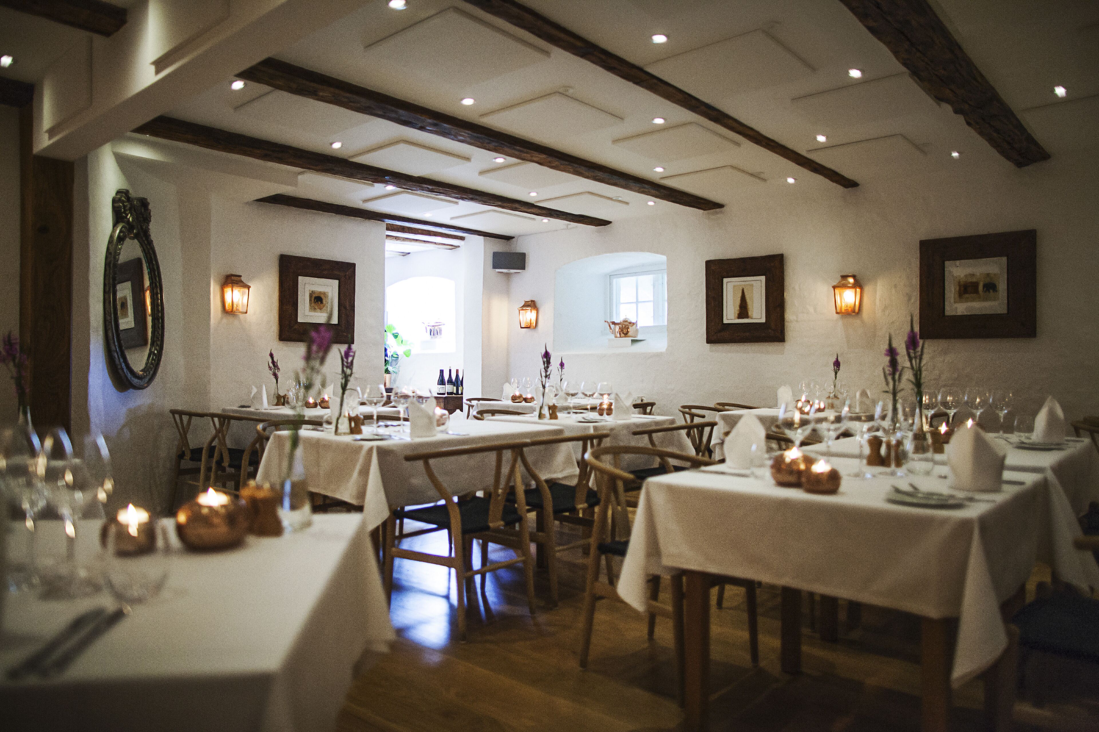 A dining area with tables set for a meal, wooden beams on the ceiling, and framed pictures on the walls.