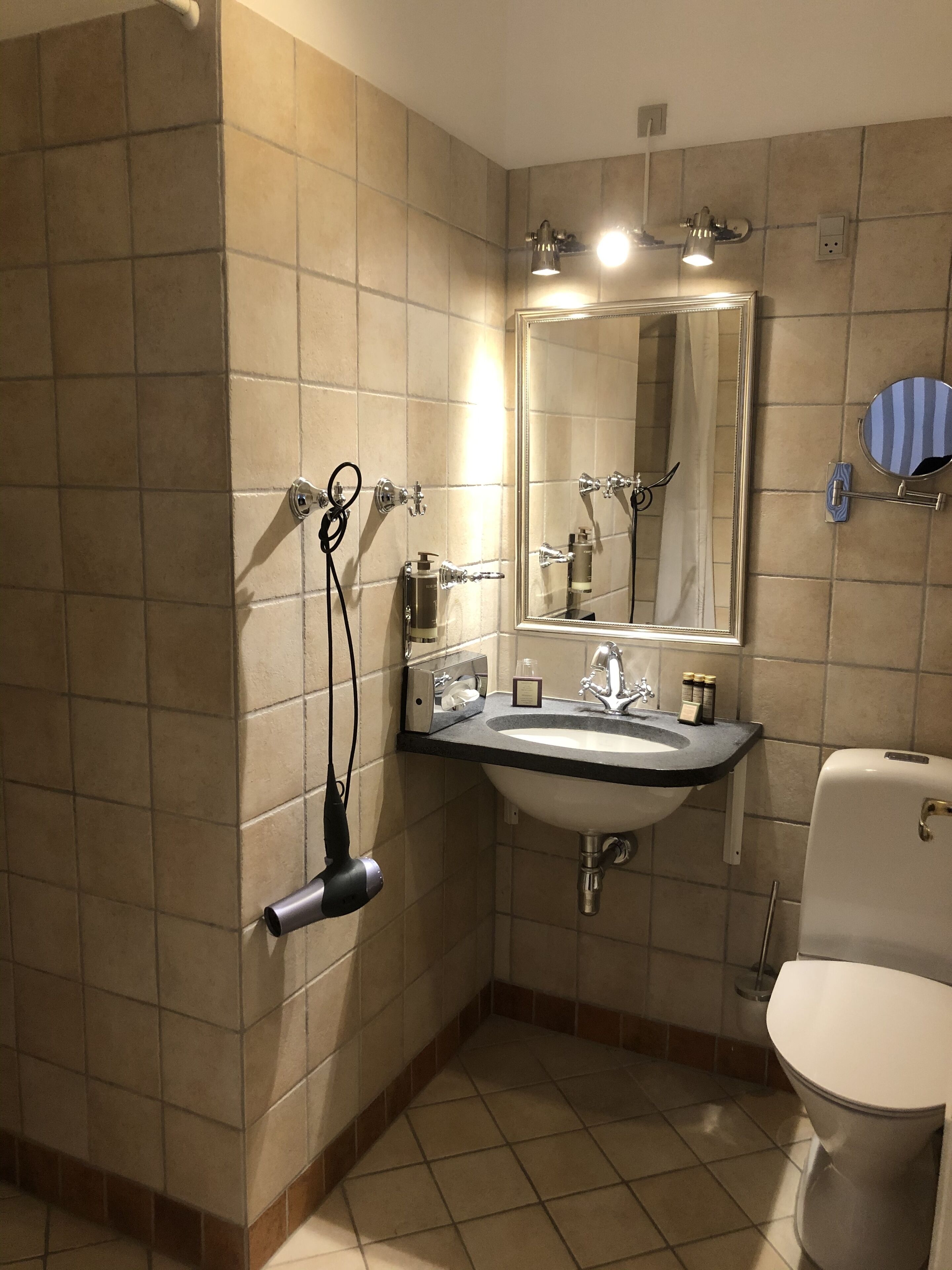 Baño con lavamanos, espejo y retrete. Hay un secador de pelo montado en la pared.