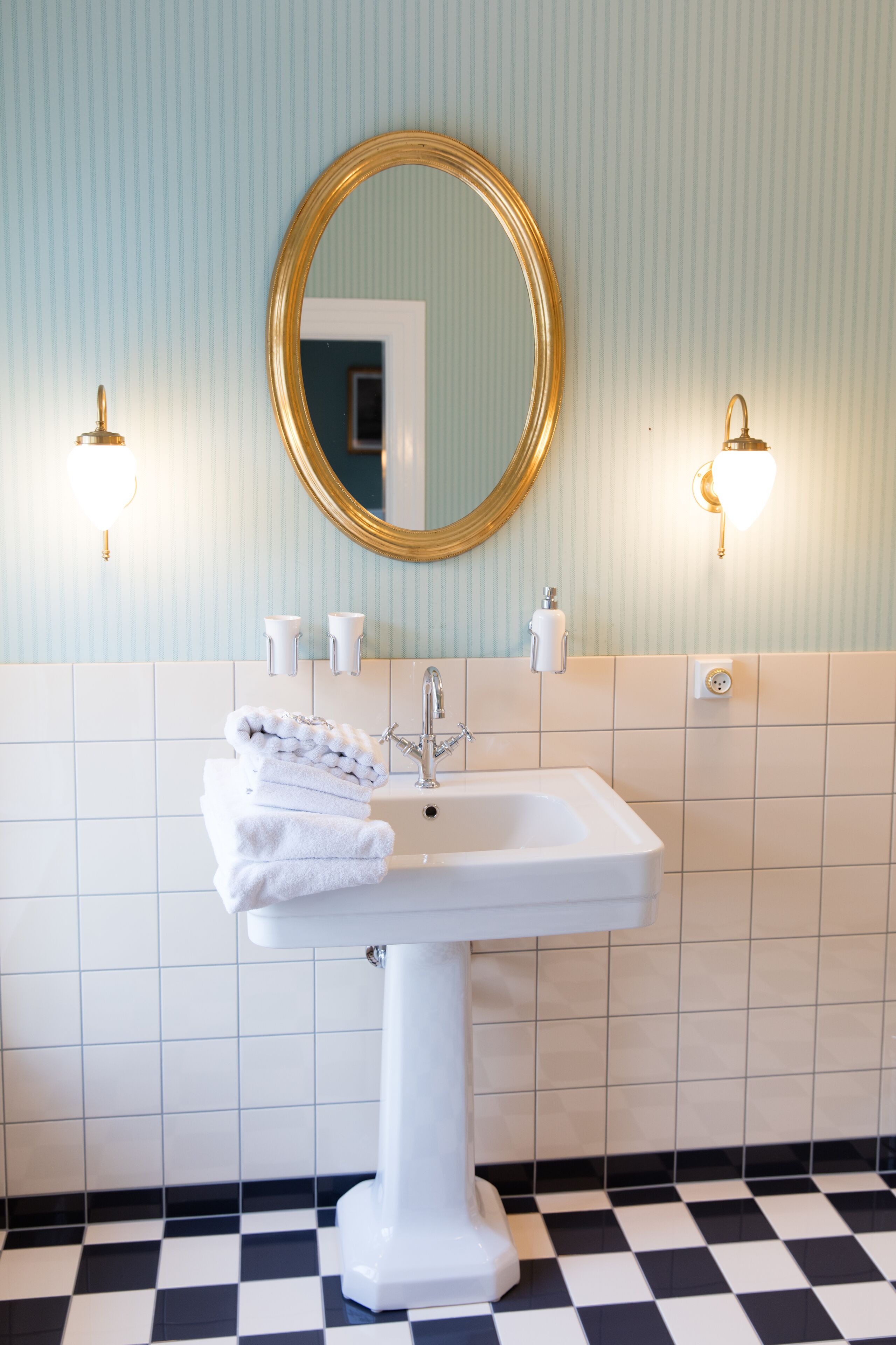 Une salle de bain avec un lavabo sur pied blanc, un miroir à cadre doré et deux appliques murales.