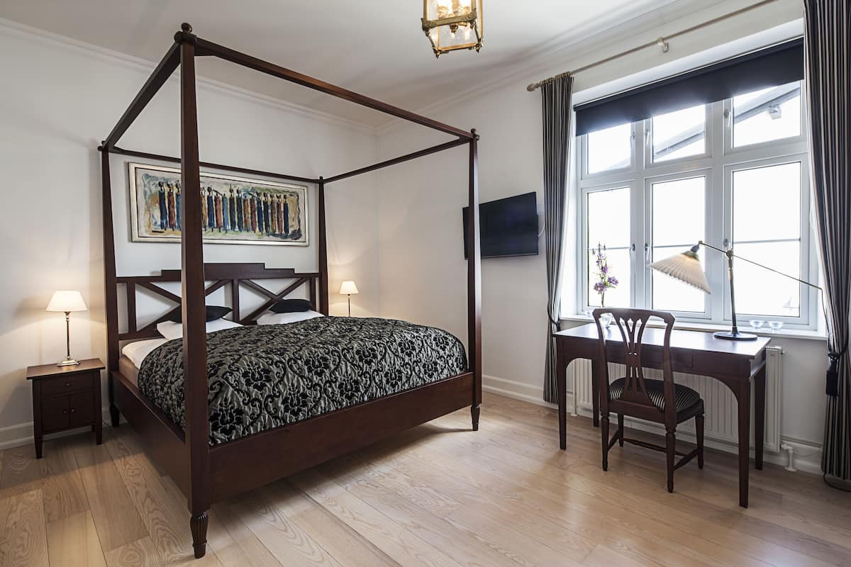 Una cama con dosel con un edredón oscuro estampado, una mesita de noche de madera con una lámpara, un escritorio con una silla y un gran ventanal con cortinas.