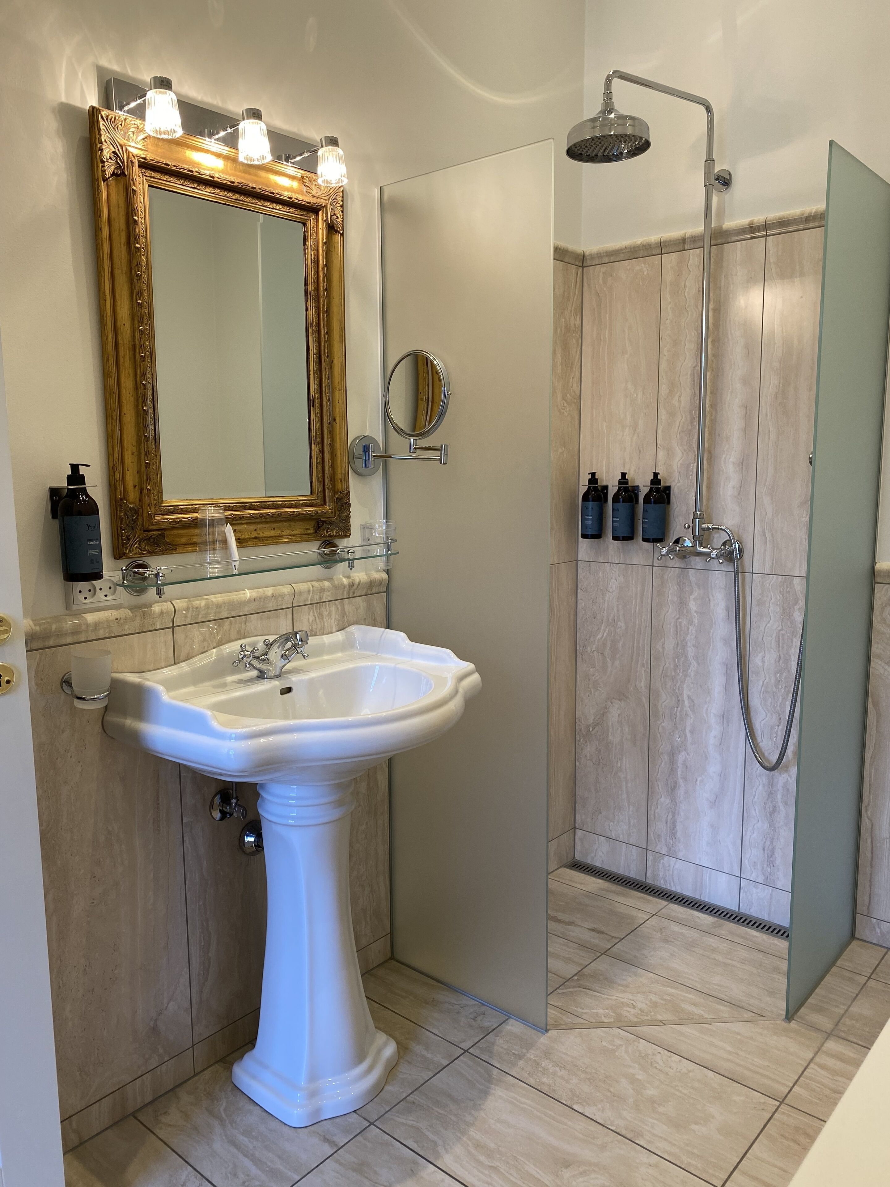 A bathroom with a pedestal sink, a large mirror with lights, a handheld showerhead, and toiletries on the wall.