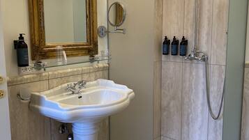 A bathroom with a pedestal sink, a large mirror with lights, a handheld showerhead, and toiletries on the wall.