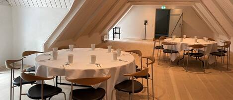 A spacious, well-lit room with multiple round tables and chairs, arranged in a semi-circular layout. The room features a sloped ceiling with wooden beams and a skylight, providing natural light.