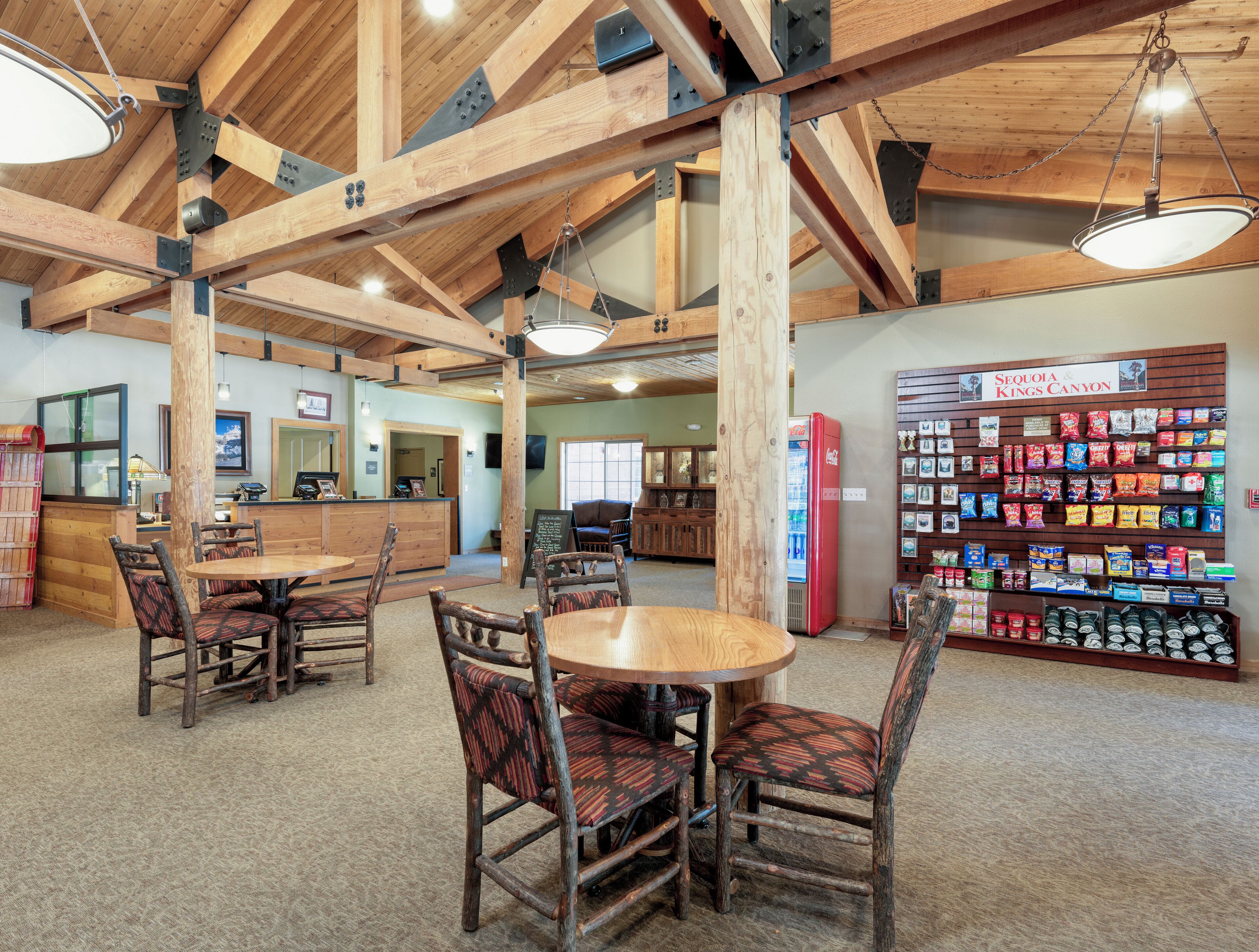 A rustic interior with wooden beams, a central round table with chairs, a Coca-Cola refrigerator, and a snack display wall.