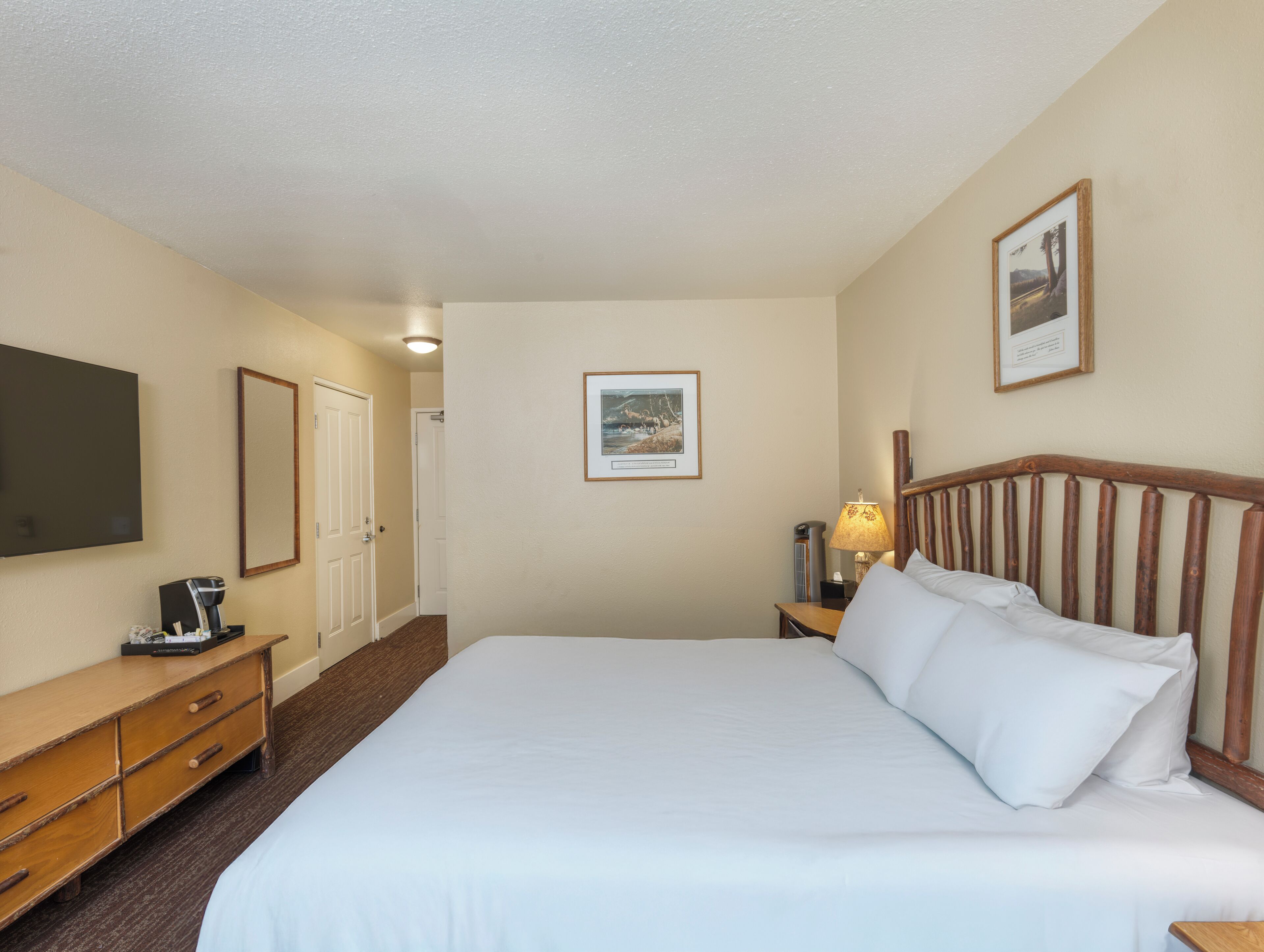 A hotel room with a large bed, a wooden headboard, a bedside table with a lamp, a TV mounted on the wall, a dresser, and a framed picture on the wall.