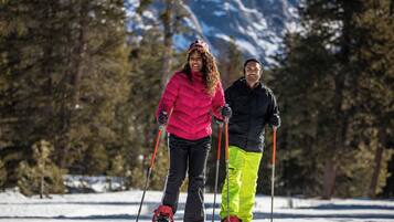 Deux personnes font du ski sur une pente enneigée, avec des arbres et des montagnes en arrière-plan.